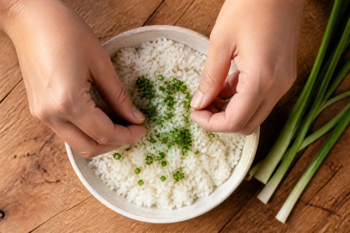 Mains disposant des herbes fraîches ciselées sur un bol de riz blanc, plan serré sur une planche de bois brut